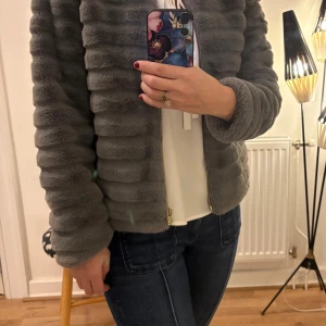 Grå pälsjacka från Pomed Lux - Säljer en fluffig grå pälsjacka från Pomed Lux med snygga horisontella ränder och guldfärgad dragkedja. Jackan är kortare i modellen och har långärmade ärmar. Insidan är fodrad med diskret prickigt tyg. Perfekt för dig som gillar mjuka och trendiga jackor.