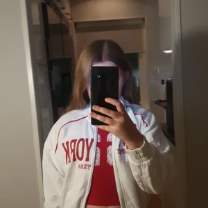 Vit sportig vindjacka med röd text - Sportig vit vindjacka med röda detaljer och texten 'NEW YORK SPORT TEAM' på bröstet. Jackan har dragkedja framtill, ribbade muddar och röda linjer längs axlarna. Perfekt för en chill och trendig streetstyle-look.