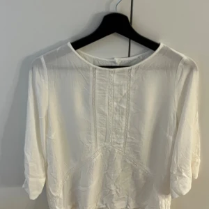 Vit blus med knappar i ryggen - Säljer en vit blus med trekvartsärm och fina spetsdetaljer framtill. Blusen har rund halsringning och knappar längs hela ryggen. Lätt och luftig känsla, perfekt för vår och sommar.