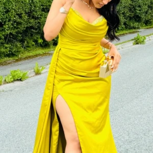 Gul långklänning med slits - Snygg gul långklänning med draperad överdel och markerad midja. Klänningen har en djup slits på sidan som ger en cool och elegant vibe. Tillverkad i ett glansigt material som faller fint och har tunna axelband. Perfekt för dig som vill sticka ut.
