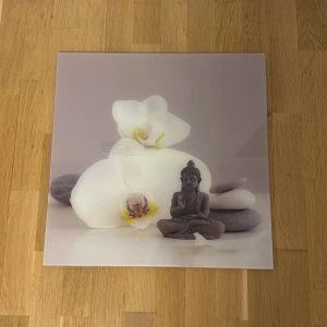 Glas tavla  - Denna produkt är en dekorativ tavla med motiv av vita orkidéer, stenar och en Buddha-staty. Tavlan har en lugn och harmonisk design, perfekt för att skapa en avslappnande atmosfär i hemmet. Skicket ser ut att vara mycket gott utan synliga skador eller slitage.