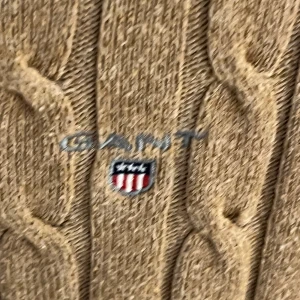 Beige kabelstickad tröja från GANT - Snygg beige kabelstickad tröja från GANT i storlek XS. Tröjan har rund hals, ribbade muddar och ett broderat GANT-logo på bröstet. Den är långärmad och har en klassisk, normal passform som sitter riktigt bra. Perfekt för dig som gillar stilrena och tidlösa plagg.