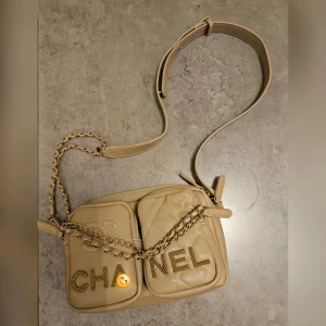 Beige axelväska från Ch*nel - Säljer en beige axelväska från Ch*nel med guldfärgad kedja och justerbar rem. Väskan har två framfickor med quiltad design och stora CH*NEL-bokstäver i guld. Perfekt för dig som vill ha en snygg och trendig väska med lyxig känsla.