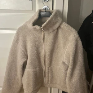 Beige teddyjacka med dragkedja - Mysig beige teddyjacka med hög krage och hel dragkedja framtill. Jackan har två stora, perfekt för att hålla värmen under kyliga dagar. Den fluffiga ytan ger en skön och trendig look.