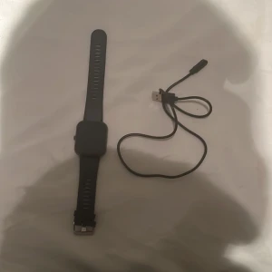 Svart smartklocka med laddkabel - Säljer en stilren svart smartklocka med rektangulär display och justerbart silikonarmband. Klockan har en metallspänne och kommer med en svart USB-laddkabel. Perfekt för dig som vill ha koll på tiden och dina aktiviteter.