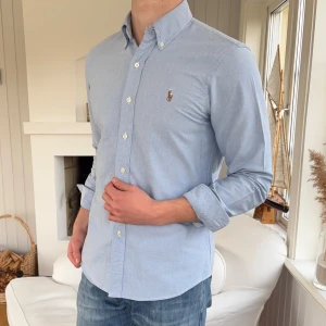 Ljusblå skjorta från Ralph Lauren - Klassisk ljusblå skjorta från Ralph Lauren med button-down krage och broderad logga på bröstet. Skjortan har lång ärm, vita knappar och är tillverkad i mjuk bomull. Perfekt för en clean och stilren look.