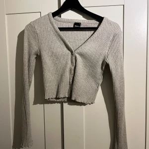 Grå ribbad croppad topp från Gina Tricot - Säljer en ljusgrå croppad topp från Gina Tricot med ribbad struktur och v-ringning. Toppen har långa ärmar, knappar framtill och vågig kant nedtill. Perfekt för dig som gillar stilrena och bekväma plagg.