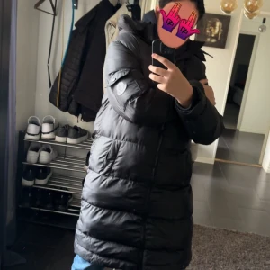 Svart lång pufferjacka Moncler - Säljer en svart lång pufferjacka från Moncler med huva och diskret logga på ärmen. Jackan har dragkedja framtill och är riktigt fluffig och varm, perfekt för kalla dagar. Passformen är normal och den har långa ärmar.