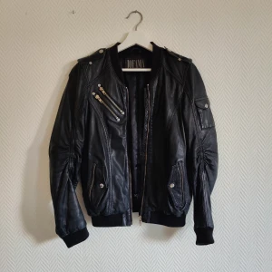 Svart skinnjacka från Jofama - Svart skinnjacka från Jofama  i bombermodell. Detaljer som dubbla dragkedjor på bröstet, ribbade muddar och axelpartier med tryckknappar. Jackan har innerficka  med dragkedja samt två sidofickor med knappstängning. Skinnet kan behöva lite omtanke och smörjning då den hängt på vinden ett tag!