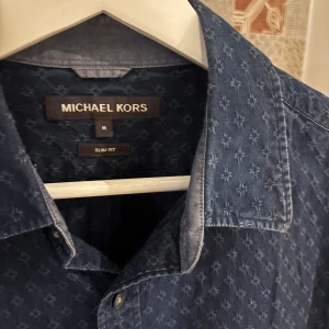 Blå slim fit skjorta Michael Kors - Snygg blå skjorta från Michael Kors i slim fit med diskret mönster och klassisk krage. Skjortan har långa ärmar och knappar framtill med märkets logga. Perfekt för dig som gillar stilrena detaljer och en modern passform.