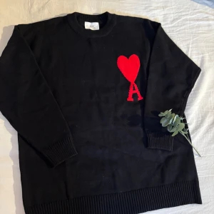 Svart stickad tröja från Ami Paris - Svart stickad tröja från Ami Paris med röd hjärt- och A-detalj på bröstet. Tröjan har rund hals, ribbade muddar och är långärmad. Perfekt för dig som gillar stilrena plagg med en twist.