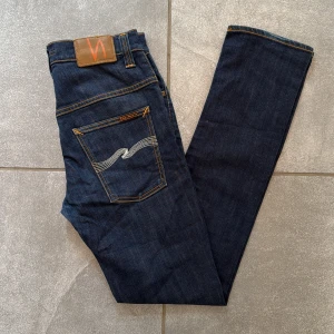 Mörkblå jeans från Nudie Jeans, W31 L34 - Snygga mörkblå jeans från Nudie Jeans med klassisk orange söm och ikonisk broderad bakficka i vitt. Modellen är Thin Finn (slimfit). Tillverkade i slitstarkt bomullsjeansmaterial, perfekta för dig som gillar stilrena och tidlösa jeans.
