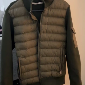 Olivgrön pufferjacka från Moncler - Snygg olivgrön pufferjacka från Moncler med quiltad front och ärmficka med logotyp. Jackan har dragkedja framtill och två sidofickor med zip. Materialet är polyester och jackan har en normal passform. Perfekt för dig som vill ha en clean och stilren look.Kan gå ner i pris eller byta mot något