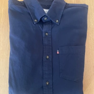 Marinblå Oxfordskjorta från Lexington S - Snygg marinblå Oxfordskjorta från Lexington med klassisk button-down krage och bröstficka med liten flaggdetalj. Skjortan har knappar framtill och är tillverkad i mjuk bomull. Perfekt för dig som gillar stilrena och tidlösa plagg.