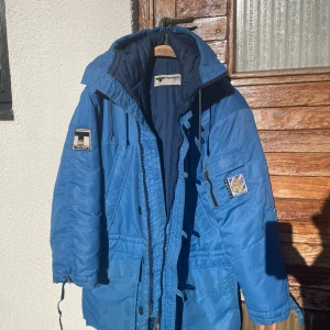 Blå parkasjacka från Tenson - Blå parkasjacka från Tenson med huva, flera fickor och coola patchar på ärmarna. Jackan har snörning i midjan och vid ärmslut, samt dragkedja framtill. Perfekt för kalla dagar med sin långa och rymliga passform.