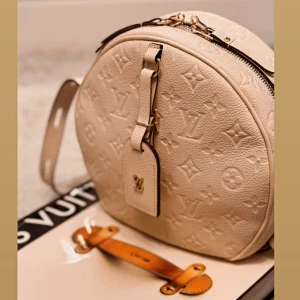 Louis Vuitton beige rund axelväska - Säljer en rund axelväska från Louis Vuitton i beige skinn med det klassiska LV-monogrammet präglat över hela väskan. Väskan har guldfärgade detaljer, dragkedja och en snygg tag med LV-logga framtill. Perfekt för dig som vill ha en unik designerbag.