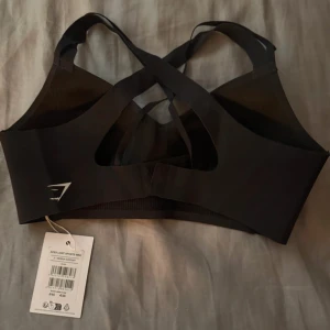 Svart sport-bh från Gymshark - Sportig svart sport-bh från Gymshark med korsade axelband i ryggen och stilren logga framtill. Modellen heter Apex Limit och har en tight passform med bred resår under bysten. Perfekt för träning och ger bra stöd. Materialet är mjukt och stretchigt syntet.