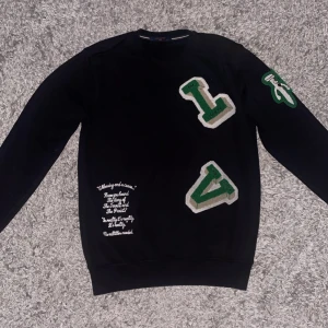Svart Louis Vuitton sweatshirt M - Svart sweatshirt från Louis Vuitton med stora gröna och vita LV-applikationer på framsidan och ärmen. Tröjan har rund hals, lång ärm och broderad text på nedre delen. .