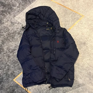 Mörkblå dunjacka Polo Ralph Lauren - Snygg mörkblå dunjacka från Polo Ralph Lauren med huva och röd broderad logga på bröstet. Jackan har dragkedja framtill, praktiska fickor med dragkedja och justerbar nederkant. Perfekt för kalla dagar och har en clean, sportig vibe.