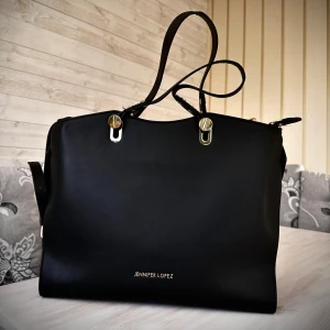 Svart handväska från Jennifer Lopez - Stilren svart handväska från Jennifer Lopez med guldiga metalldetaljer och dubbla handtag. Finns en längre rem att sätta fast och ha över ena axeln också. Köptes runt ca 1200 kronor💕I gott skick!