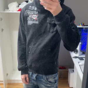 Svart hoodie Geographical Norway - Svart hoodie från Geographical Norway med tryck och broderade detaljer fram, bak och på ärmen. Stor norsk flagga på ryggen, bröstficka med knapp och känguruficka. Mjuk insida och huva med grått foder. Storlek L men sitter som M/S Jag är 181 72kg