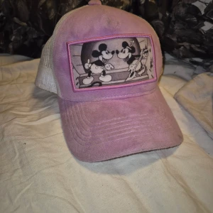 Ljuslila Disney keps med mesh - Säljer en ljuslila keps med vit mesh på sidorna och baksidan. Framtill finns en broderad patch med klassiskt svartvitt motiv av Musse och Mimmi Pigg. Kepsen har böjd skärm och cool streetstil, perfekt för dig som gillar retro och Disney.