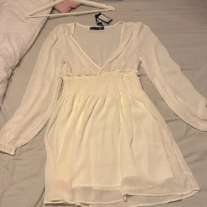 Vit omlottklänning från 157 XS - Superfin vit kortklänning från 157 med omlott-v-ringning, långa ärmar och smockad midja. Klänningen har volangdetaljer vid midjan och är tillverkad i ett lätt, luftigt tyg som ger en flowy känsla. Perfekt för dig som gillar en romantisk och stilren look.