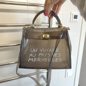 Väska - Un Voyage Bag, använd 1 gång. Väskan är genomskinlig med ett långt axelremsband och ett handtag. 