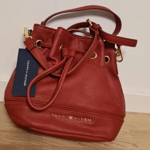 Oanvänd fin Tommy Hilfiger väska - Oanvänd fin Tommy Hilfiger väska i röd färg. Gulddetaljer. Väskan är oanvänd med prislapp på. 100% äkta och köpt i Usa   Perfekt som present eller julklapp 