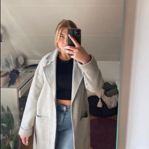 Grå kappa - Helt ny, superfin grå kappa (ull). Skick är som ny! Den har ett midjeband och två fickor. Köpt för 1000kr!