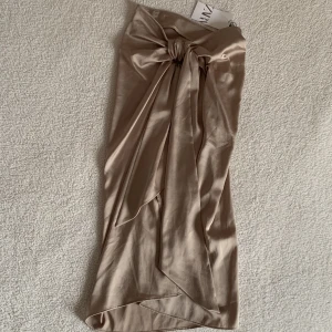 ZARA glansig guldig / beige midikjol med släp - Den här kjolen är magisk! Färgen är mer åt det guldiga hållet, men var svårt att fånga på bild. Tyvärr inte kommit till användning. Nyskick med tags kvar. 🌻Dold dragkedja på sidan. Stilig knytdetalj med långt hängande släp.