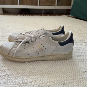Vita Adidas Stan Smith sneakers - Ett par vita adidas Stan Smith sneakers med mörk blå häl. De har en del märken i materialet men mest på insidan av foten, så inte så synliga när man har dom på.