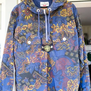 Stone Island X Supreme Anorak  - Nylon metall 5C Anorak Marinblå  5-färgstryckt med ett motiv tillverkat av Supreme exklusivt för detta samarbete. Modellen är återutgåvan av en av de första Stone Island-kollektionenerna '982. Den har Badge som hålls av metallknappar Supreme tryck baktill