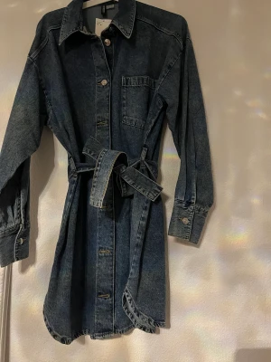 Denim klänning - En jättefin denimklänning med silverdetaljer och knappar. Superfin och aldrig använd, prislappen hänger kvar! Man får även med ett band som man kan knyta i midjan! Syns inte i bilden men den har en skjort-krage! 