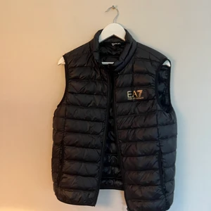 Emporio Armani EA7 väst strl. L svart - En svart väst från Emporio Armani EA7 i svart med guldiga tryck. Oanvänd helt i nyskick. Den är mindre i storleken men passar jättebra om du har M eller om du vill ha en lösare form och har S. Köparen betalar frakten.