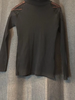 Svart polotröja med meshdetaljer - Snygg svart polotröja med lång ärm och meshpartier med små prickar över axlarna. Tröjan är ribbad och har en tight passform som ger en modern look. Perfekt för dig som gillar stilrena plagg med en twist.