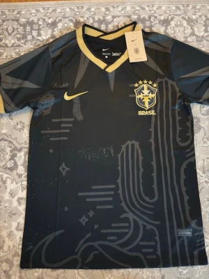 Svart Brasil Nike fotbollströja - Snygg svart fotbollströja från Nike med Brasil-logga och gula detaljer. Tröjan har ett unikt grafiskt mönster, gul V-ringad krage och Nike Swoosh på bröstet. Perfekt för dig som vill sticka ut på planen eller heja på Brasilien.