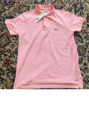 Rosa pikétröja från Lacoste - Klassisk rosa pikétröja från Lacoste med korta ärmar och krage. Tröjan har knappar framtill och den ikoniska krokodilloggan på bröstet. Perfekt för en avslappnad och stilren look.