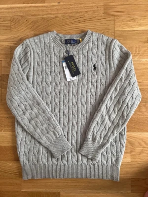 Polo Ralph Lauren Sweater  - Snygg grå kabelstickad tröja från Polo Ralph Lauren med rund halsringning och klassisk broderad logga på bröstet.