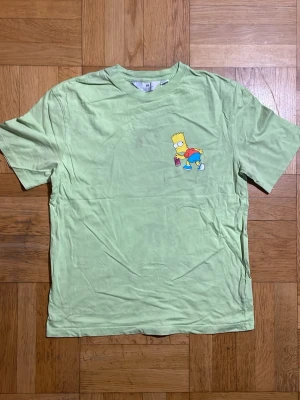 Grön Bart Simpson t-shirt - Ljusgrön t-shirt med tryck av Bart Simpson som sprayar graffiti och texten 'SORRY OR WHATEVER' på ryggen. Klassisk passform och korta ärmar, perfekt för dig som gillar The Simpsons och streetstyle.