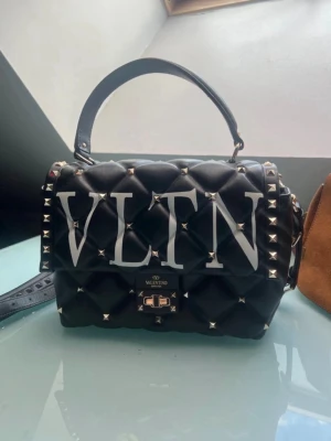 Svart Candystud Valentino Garavani väska - Snygg svart handväska från Valentino med stora VLTN-bokstäver framtill. Väskan är quiltad i mjukt läder och dekorerad med silverfärgade nitar. Har både handtag och axelrem samt ett praktiskt ytterfack på baksidan. Perfekt för dig som gillar edgy och lyxig stil. Ny och i jätte jätte fint skick