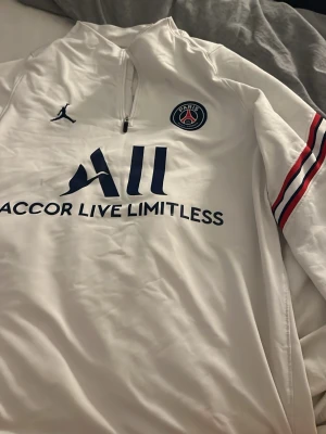 Vit PSG träningströja Jordan - Snygg vit träningströja från PSG och Jordan med halv dragkedja, klubbmärke på bröstet och blå text. Röd, vit och blå rand på ärmen samt PARIS-tryck på ena ärmen. Perfekt för fotbollsträning eller chill. Tillverkad i lätt och funktionellt material. Klippt nedtill men syns ej så mycket 