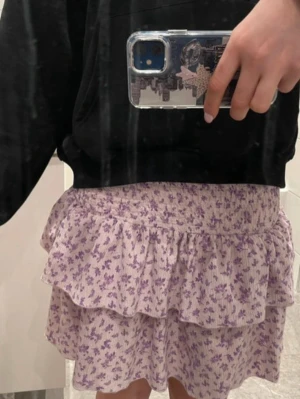 Lila volangkjol med blommor från gina tricot - Superfin volangkjol med två lager och smock i midjan som gör den både bekväm och följsam. Kjolen har ett mjukt, ljuslila tyg med små lila blommor – perfekt till både vardag och fest. Sitter fint, ger en lätt och flowy känsla och passar lika bra med hoodie som med en enkel topp. Midjemåttet är 52 cm och i är i storlek xxs. Men pågrund av resåren passar den även större storlekar.