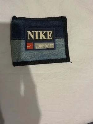 Nike denim plånbok med logga - Säljer en snygg plånbok från Nike i blå denim med svart kant och tydlig Nike-logga samt texten 'Just Do It' på framsidan. Plånboken har flera fack för kort och sedlar samt ett fack med dragkedja. Perfekt för dig som gillar sportig och avslappnad stil.
