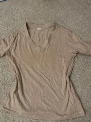 Beige v-ringad topp i bomull - En enkel beige topp med v-ringning och korta ärmar. Tillverkad i mjuk bomull, perfekt för en avslappnad och stilren look. Passformen är normal och tyget är tunt och skönt mot huden.