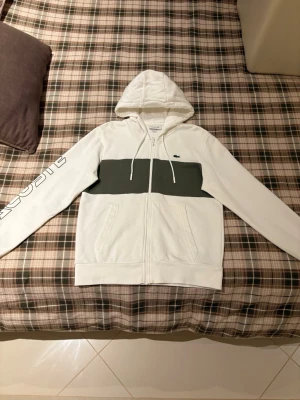 Vit hoodie från Lacoste med tryck - Snygg vit hoodie från Lacoste med dragkedja, huva med snören och en bred mörkgrön rand över bröstet. På vänster ärm står det 'LACOSTE' i stora bokstäver. Klassisk krokodil-logga på bröstet och två fickor framtill. Perfekt för en chill och sportig look.
