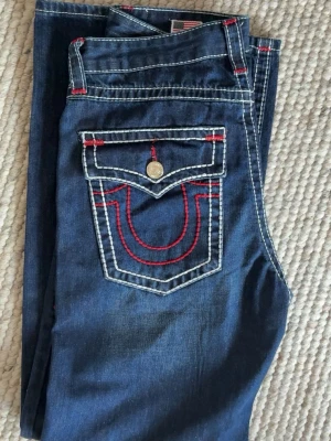 True Religion jeans - blåa jeans från True Religion med vita och röda kontrastsömmar. Klassisk femficksmodell med raka ben och dekorativ ficka bak med lock och knapp. Snygga detaljer och ikonisk logga på bakfickan. Jag är 185cm lång och väger 70kg och dessa jeans har en mer lose fit på mig vilket syns i bilderna.