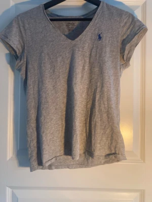Grå v-ringad t-shirt från Polo Ralph Lauren - Snygg grå t-shirt från Polo Ralph Lauren med v-ringning och korta ärmar. Klassisk blå logga broderad på bröstet. Tillverkad i mjuk bomull som känns skön mot huden. Perfekt basplagg till garderoben.