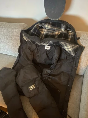 Svart dunväst från Burberry - Säljer en svart dunväst från Burberry med klassiskt rutigt foder på insidan. Västen har dragkedja och tryckknappar samt en snygg patch med Burberry London England på ena sidan. Perfekt för lager-på-lager och riktigt stilren.