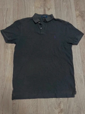 Ralph Lauren pikétröja - Snygg och tidlös pikétröja från Ralph Lauren i svart färg med klassisk lila broderad logga på bröstet. Perfekt både till vardag och lite mer uppklätt.  ✔️ Modell: Custom Fit ✔️ Storlek: M ✔️ Färg: Grå ✔️ Skick: Bra skick – normalt använd, inga större defekter ✔️ Material: Mjuk bomullspiké  Passar lika bra till jeans som chinos. En riktig staple i garderoben!
